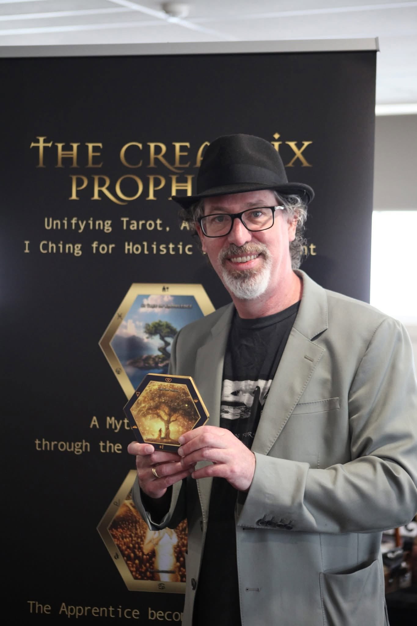 Benjamin Rose smiling and holding a gold tree hexagonal Creatrix card in front of The Creatrix Prophecy banner.